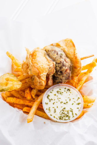 The Deep Fried Bacon Burger Dog Slider on a Stick is served with ranch dressing, fries, and a fried pickle. 