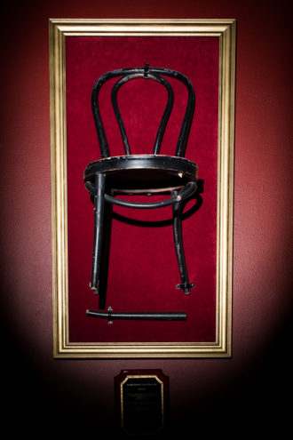 FOUR DAY WEEKEND’S ORIGINAL STAGE CHAIR: It hangs in the theater, and a plaque beneath it reads: “This chair was adopted (stolen) from Casa Mañana’s Theater on the Square on Four Day Weekend’s opening night, February 28, 1997. It has been with us ever since. This chair has appeared in more than 4,000 shows and 25,000 scenes. It has and always will be an original member of the cast. This beloved chair has been a marching band drum, hooker’s high heel shoe, crown, canoe, car, airplane, tank, toilet, couch, hospital bed, bagpipe, prison cell, bird cage, angel wing, and countless other memories. Our dependable black chair met its untimely demise in an unfortunate bobsledding accident on January 17, 2015. You are gone but not forgotten. Rest in pieces, old friend.”