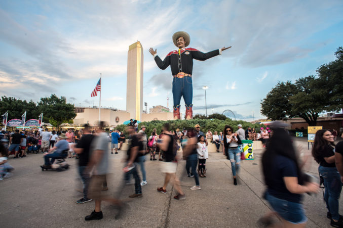 An image of Fair Park during the State Fair of Texas in 2018, with Big Tex in the center and fair-goers walking around.