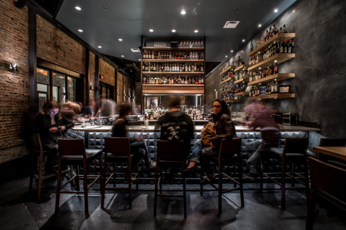 The interior of a bar with people sitting at the bar with bottle of liquor in the background.