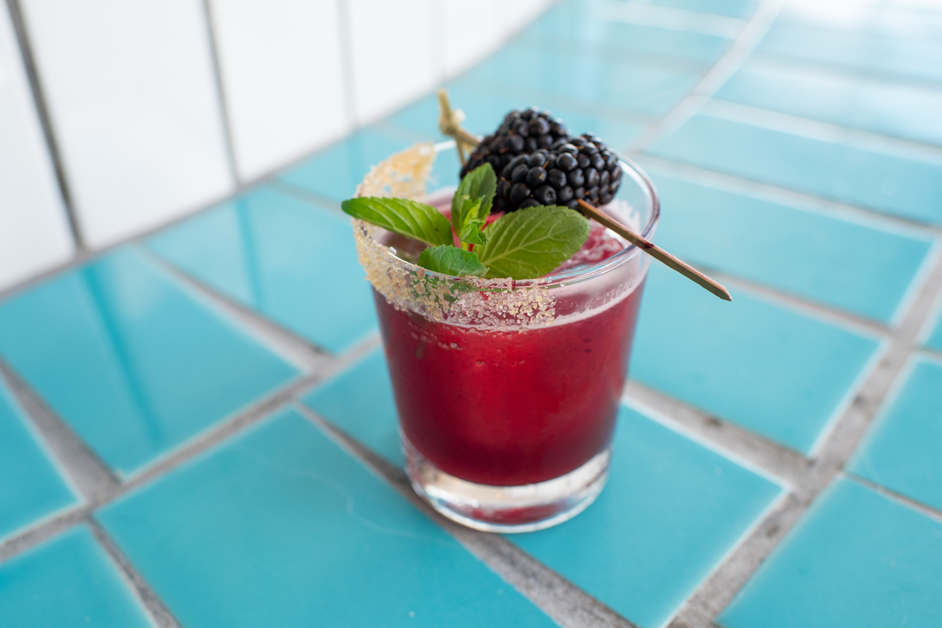A blackberry tequila cocktail with berry and mint garnish on a blue tile counter.