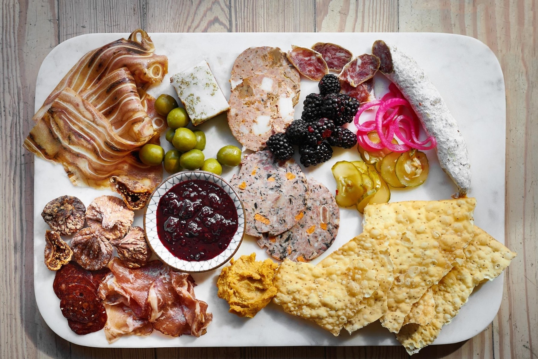 A packed charcuterie board topped with green olives, large seedy crackers, pickles, salumi, and other various cured meats.