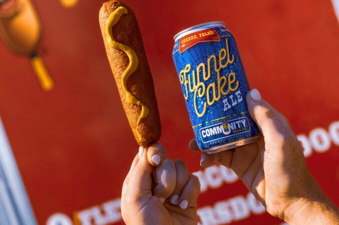 In one hand someone's holding a corn dog with mustard on it, and in the other hand a Funnel Cake ale.
