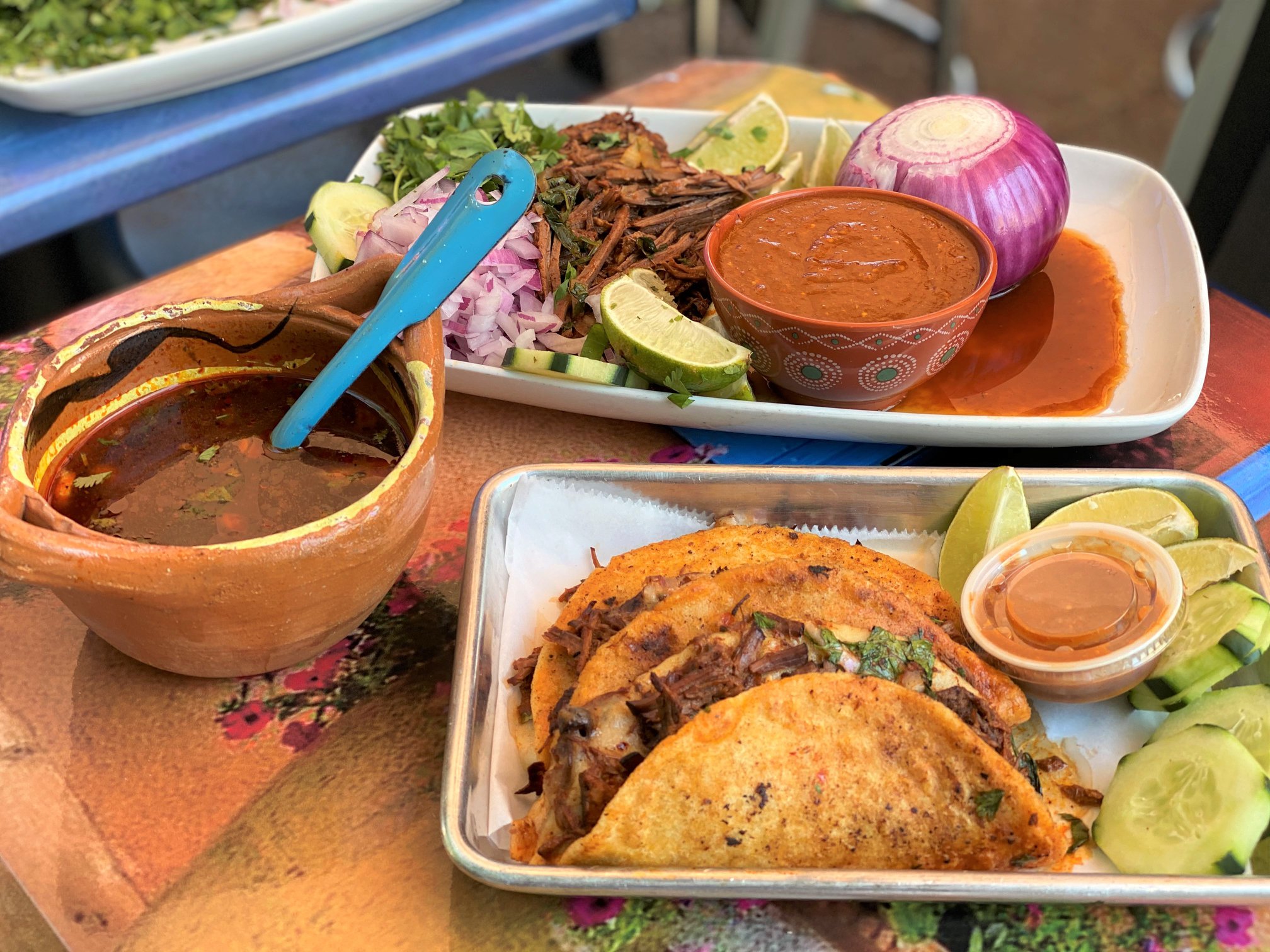 A tablescape of tacos: a tray with taco fixins, salsa, veggies; another tray with quesabirra tacos with a side of dipping stew.