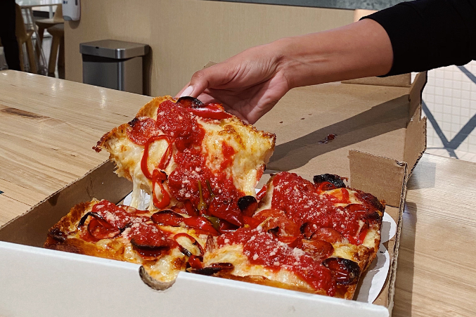 A square slice of pizza in a takeout box getting its cheese stretched away from the whole pie.