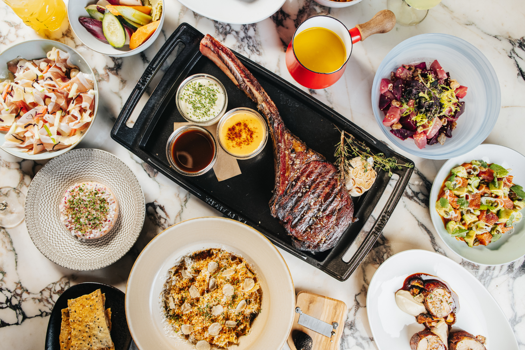 A spread of food on a table with a big tomahawk shop at the center, surrounded by sides and drinks.