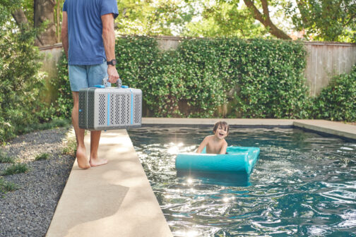 A man carrying a portable Nomad grill and walking beside a pool with a child swimming and playing.
