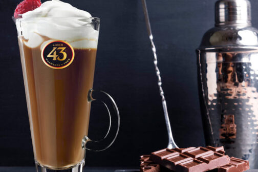 A glass of of a coffee cocktail, the carajillo, topped with whipped cream, a raspberry, next to chocolate and a cocktail shaker.