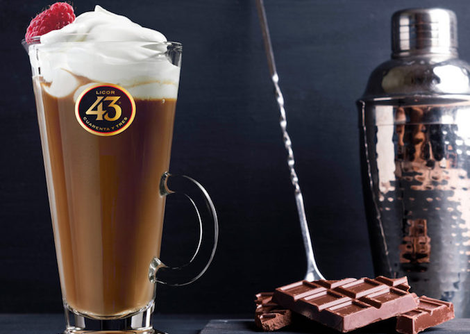 A glass of of a coffee cocktail, the carajillo, topped with whipped cream, a raspberry, next to chocolate and a cocktail shaker.