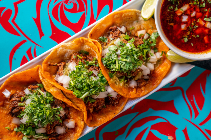 A platter of four tacos with a side of birria de res dipping sauce.