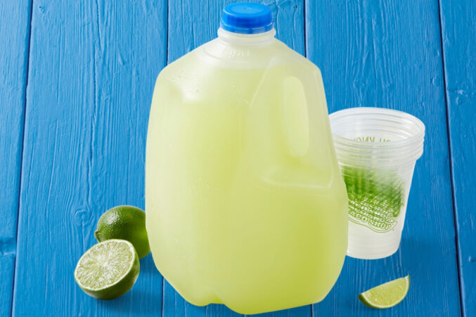 A gallon jug of margarita with plastic cups and slices of lime.