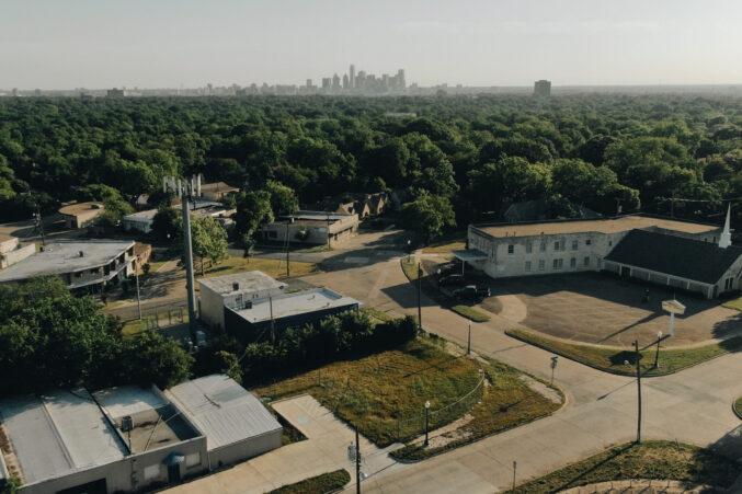 A bird's eye drone shot of the Elmwood Farm plot of land before it was turned into a working farm. There's a small street, a few residential buildings, and a patchy square of grass where the farm will eventually go.