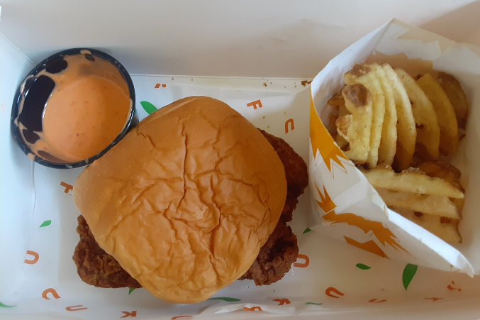 A to-go box with waffle fries, a fried chicken sandwich, and spicy mayo.