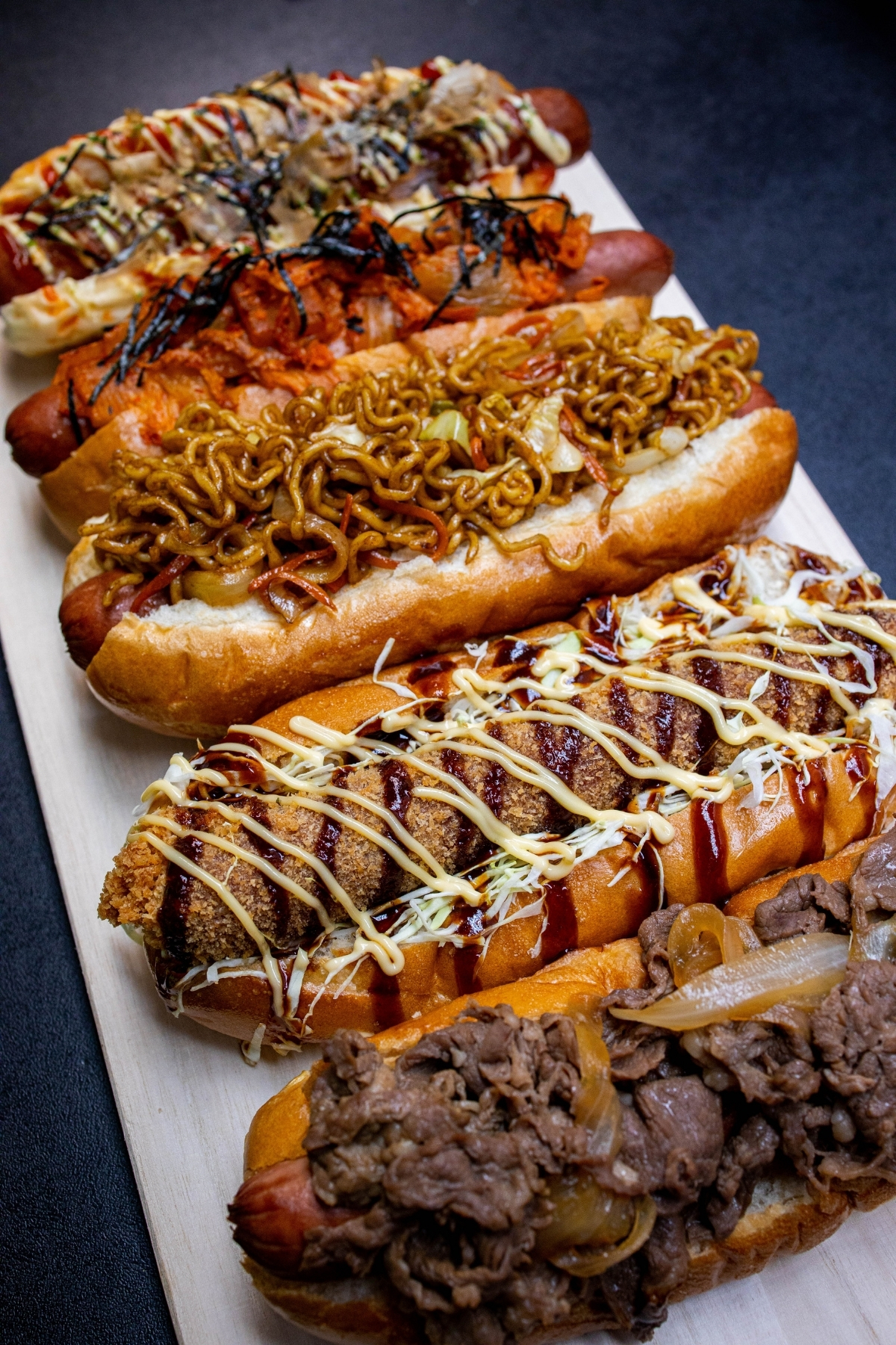 A row of Japanese-style hot dogs all spruced with a whole manner of toppings from fried tofu to yakisoba.