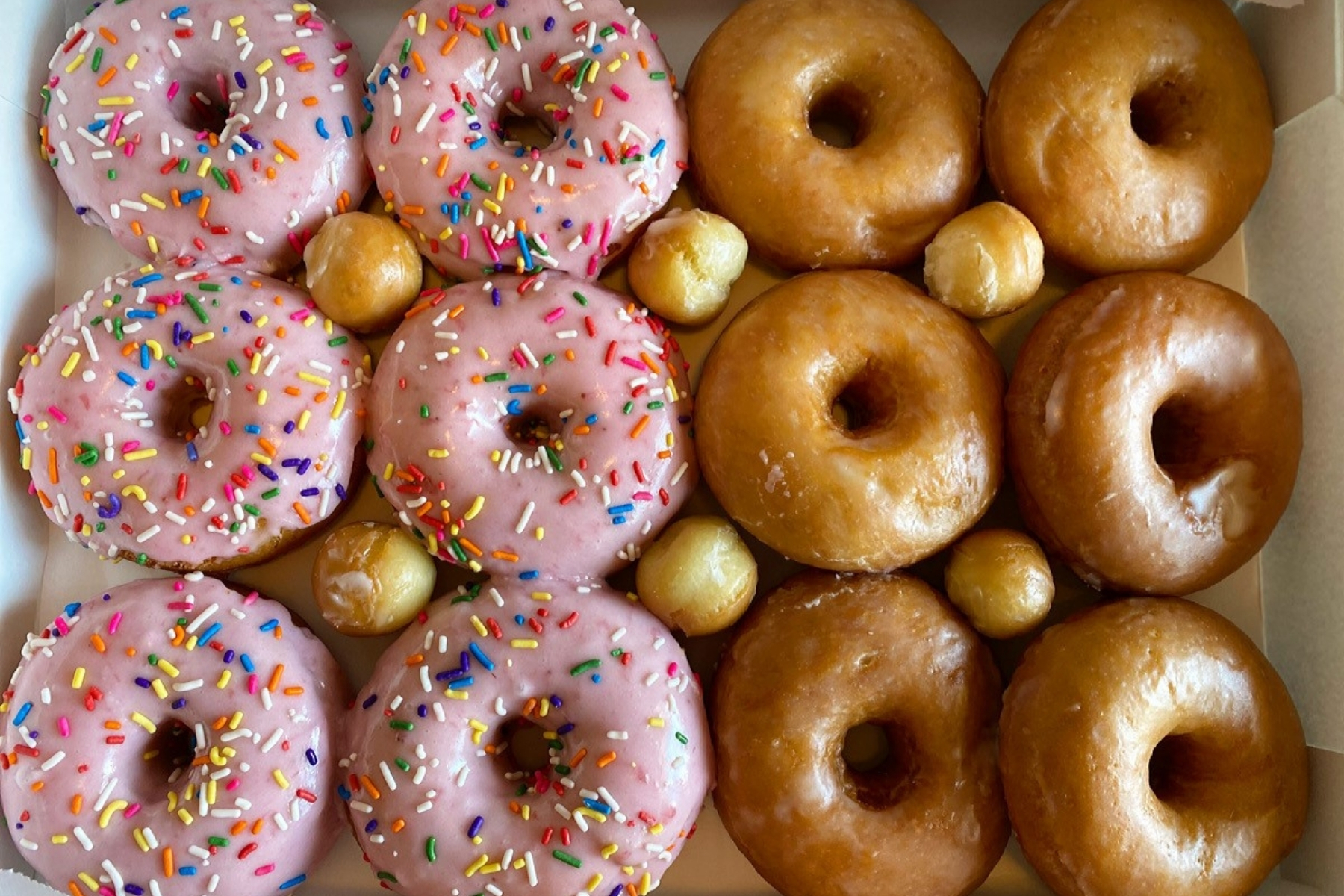 A box of vegan doughnuts. A half dozen pink doughnuts with sprinkles, and the other half dozen are plain glazed.