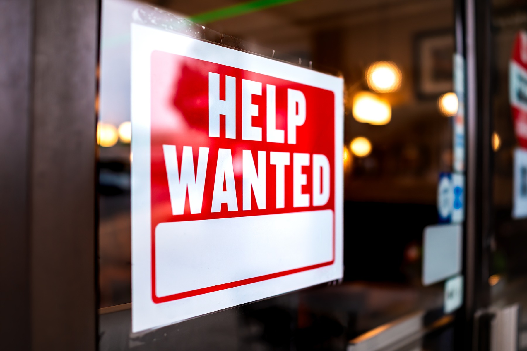 Sign text closeup for help wanted with red and white colors by entrance to store shop business building during corona virus covid 19 pandemic