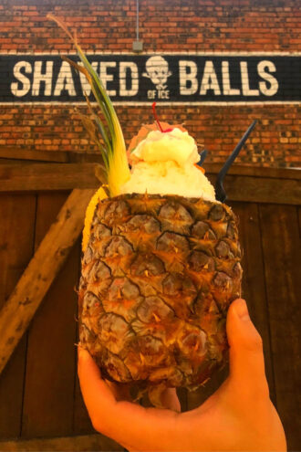 A hand holds up a a hallowed out pineapple that's filled with vanilla ice cream, shaved ice covered in pineapple and coconut syrups. And on top there's whipped cream and a maraschino cherry and tiny umbrella.