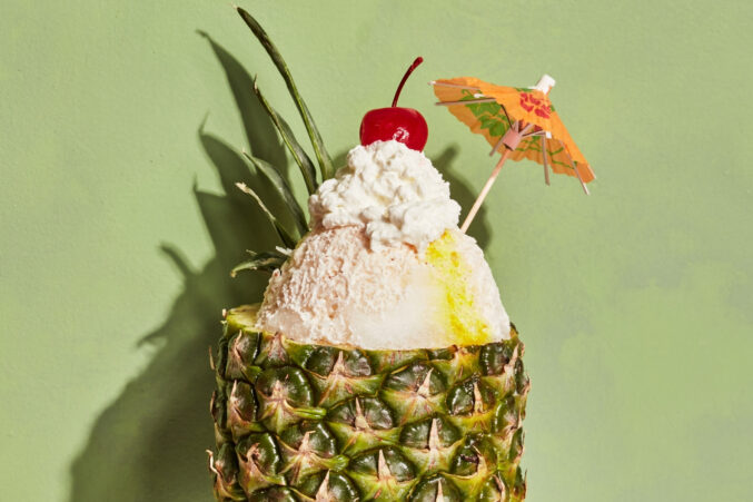 Light green background with a pineapple that’s been cut and hollowed out. Inside is coconut and pineapple shaved ice with whipped cream and cherry on top, a cocktail umbrella.