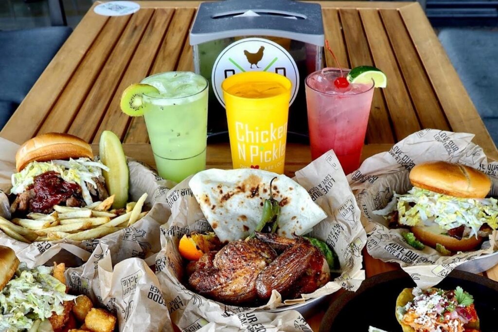 a picnic table with three drinks, burgers and fries, chicken sandwiches, and wood-fired rotisserie chicken