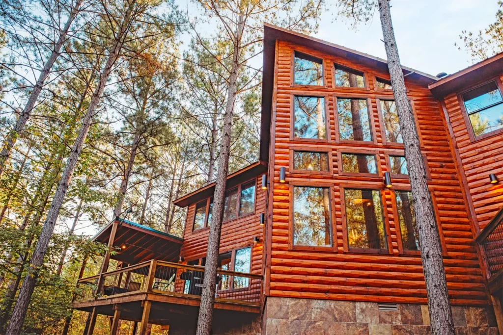 A rustic, modern cabin surrounded by tall pine trees in Beavers Bend Cabin Country, Oklahoma. The cabin features large glass windows reflecting the forest and a wooden deck with a cozy, elevated design, perfect for a relaxing getaway.