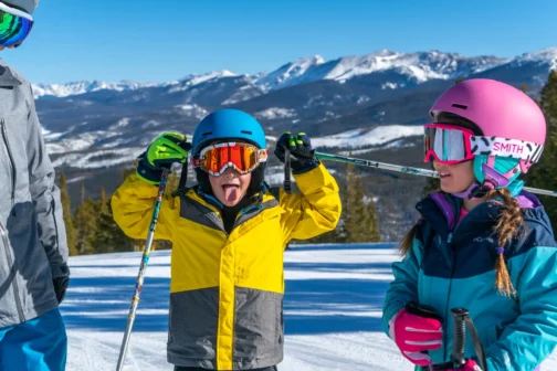 Two children in colorful ski gear enjoying a sunny day on the slopes at Winter Park Resort, Colorado. The boy in a yellow jacket playfully sticks his tongue out, while the girl in a pink helmet smiles with snow-covered mountains in the background.