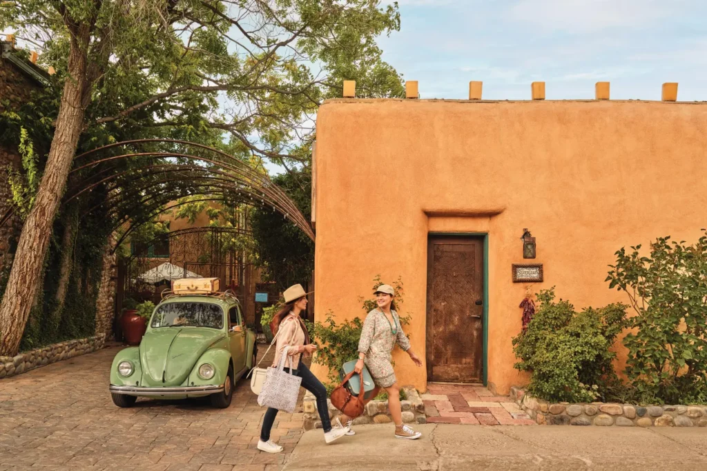 Two women walking past an adobe-style building in Santa Fe, New Mexico, with a vintage green Volkswagen Beetle parked nearby under a decorative archway surrounded by trees and greenery. The women are carrying bags and wearing casual clothing, including hats, while smiling and enjoying the sunny day.