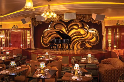 At Delilah in the Dallas Design District, an elegant, dimly lit jazz club featuring a black grand piano on a wooden stage backed by a large, ornate gold shell sculpture. Arranged beneath warm chandeliers are white-clothed dining tables with illustrated lamps and plush velvet seating.