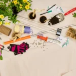 Flat lay of various gardening supplies including potted yellow and pink flowers, seedlings, hand tools, gloves, seed packets, and grow lights arranged on a light-colored paper surface. An assortment of gardening gear like twine, pruners, and soil scoops are scattered around the plants.