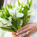 A person in a lace wedding dress holds a bridal bouquet of white lily of the valley and sweet peas. Their hand features a light pink manicure and a gold engagement ring with three diamonds.