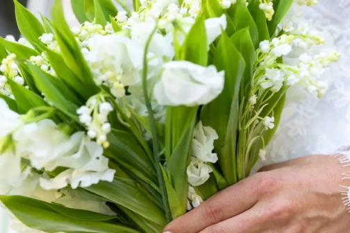 A person in a lace wedding dress holds a bridal bouquet of white lily of the valley and sweet peas. Their hand features a light pink manicure and a gold engagement ring with three diamonds.