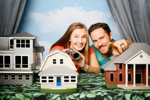 Hannah and Matthew Sams peer over several detailed architectural house models. The woman holds binoculars while the man points toward one of the houses against a blue sky backdrop.