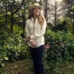 A woman with long wavy hair and a cowboy hat smiles while standing in a lush, wooded garden. She wears a cream-colored button-down shirt and a bolo tie, surrounded by green foliage and pink wildflowers.
