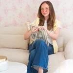 A smiling woman sits on a cream-colored bouclé sofa, working on a needlepoint project in a wooden frame. She is wearing a white t-shirt, blue jeans, and a yellow sweater draped over her shoulders against a background of pink floral wallpaper.