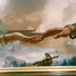 A close-up shot of a man's and woman's hands reaching toward each other, with their fingertips nearly touching in front of a framed landscape painting. The woman wears a gold charm bracelet and an engagement ring, while the man wears a suit with a silver watch.