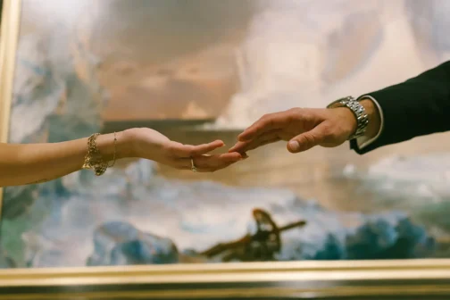 A close-up shot of a man's and woman's hands reaching toward each other, with their fingertips nearly touching in front of a framed landscape painting. The woman wears a gold charm bracelet and an engagement ring, while the man wears a suit with a silver watch.