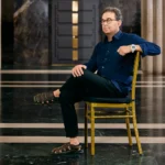 A man with glasses and a blue button-down shirt sits cross-legged on a gold chair in a large hall with marble floors and columns. He wears sandals and a white watch, looking off to the side with a neutral expression.
