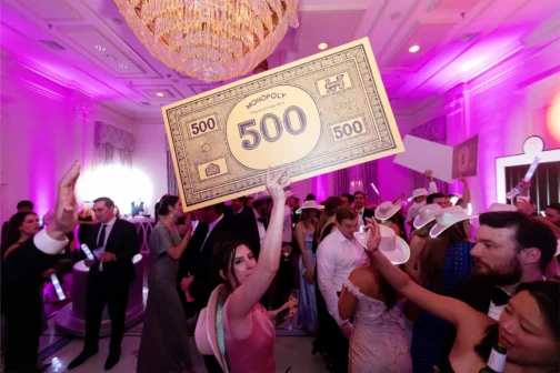A woman in a pink dress holds up a giant oversized 500 Monopoly bill above a crowded dance floor at a formal event with pink lighting and a crystal chandelier. Many guests wear western-style hats and formal attire while celebrating in the ballroom.