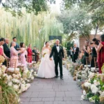 A bride in a white strapless gown and a groom in a black tuxedo walk down a stone aisle, smiling as guests applaud from rows of gold chairs. The outdoor wedding ceremony is decorated with large floral arrangements of white and pink hydrangeas under a canopy of lush green trees.