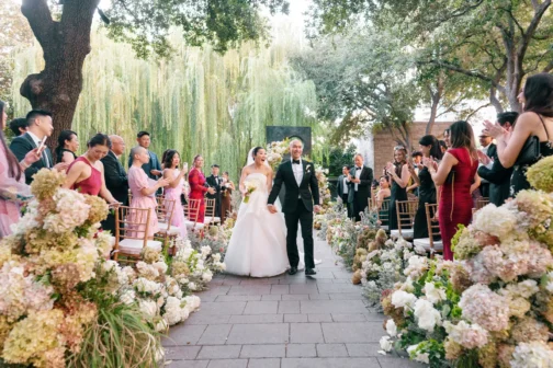 A bride in a white strapless gown and a groom in a black tuxedo walk down a stone aisle, smiling as guests applaud from rows of gold chairs. The outdoor wedding ceremony is decorated with large floral arrangements of white and pink hydrangeas under a canopy of lush green trees.