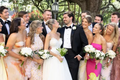 A bride in a white lace gown and a groom in a black tuxedo smile at each other, surrounded by a wedding party of men in tuxedos and women in colorful floral dresses. The group is outdoors under lush green trees, with the bridesmaids holding white bouquets.