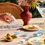 A hand wearing statement gemstone rings holds a Mahjong tile over a table set with blue tiles, a martini, olives, and cookies. The scene features elegant decor including a carved red vase and patterned linens.