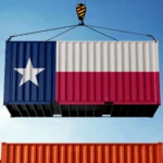 A shipping container painted with the Texas state flag is suspended in the air by a crane hook against a clear blue sky. Other colorful containers are visible at the bottom of the frame, suggesting a busy shipping port.