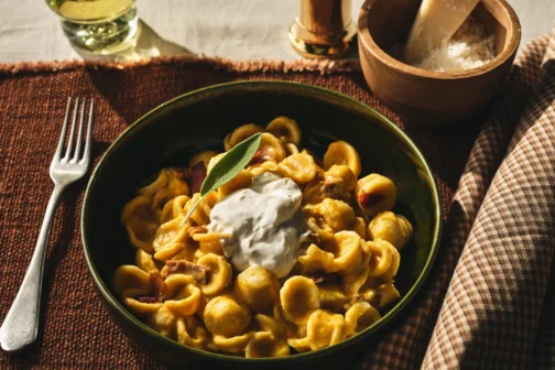 Orecchiette pasta tossed in a creamy orange sauce with pancetta, topped with a dollop of burrata cheese and a fresh sage leaf. The dish is served in a dark green bowl on a rustic brown placemat next to a fork and a bowl of grated parmesan.