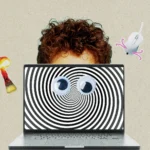A person with curly brown hair peers over the top of a laptop screen, which displays a black and white hypnotic spiral. Two large googly eyes and a computer mouse with pink squiggly legs are superimposed over the spiral, creating a surreal face.