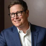 A smiling man with short brown hair and thick black-rimmed glasses, wearing a blue blazer over a white button-down shirt.