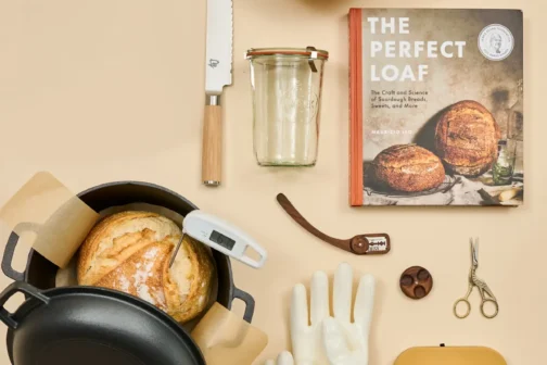 A top-down collection of sourdough baking tools including a cast iron dutch oven with a loaf inside, proofing baskets, a digital scale, heat-resistant gloves, and a cookbook titled 