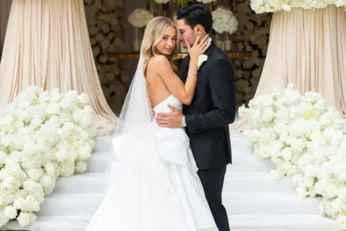 A bride in a strapless white gown and veil embraces a groom in a black tuxedo against a backdrop of white roses and draped cream fabric. The couple is posed intimately, with the bride looking toward the camera and the groom leaning his head toward hers.