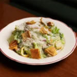 A Caesar salad on a white plate with a red rim, topped with a heavy dusting of grated parmesan cheese and large, square pita chips. The salad is served on a dark wood table.